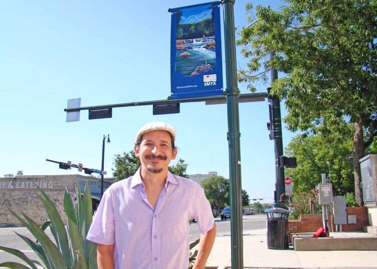 San Marcos artist Rene Perez smiles in front of a newly-installed banner downtown with his painting of a bobcat and a rattlesnake floating on the San Marcos River. Perez wanted to incorporate the city’s two school mascots in a painting.